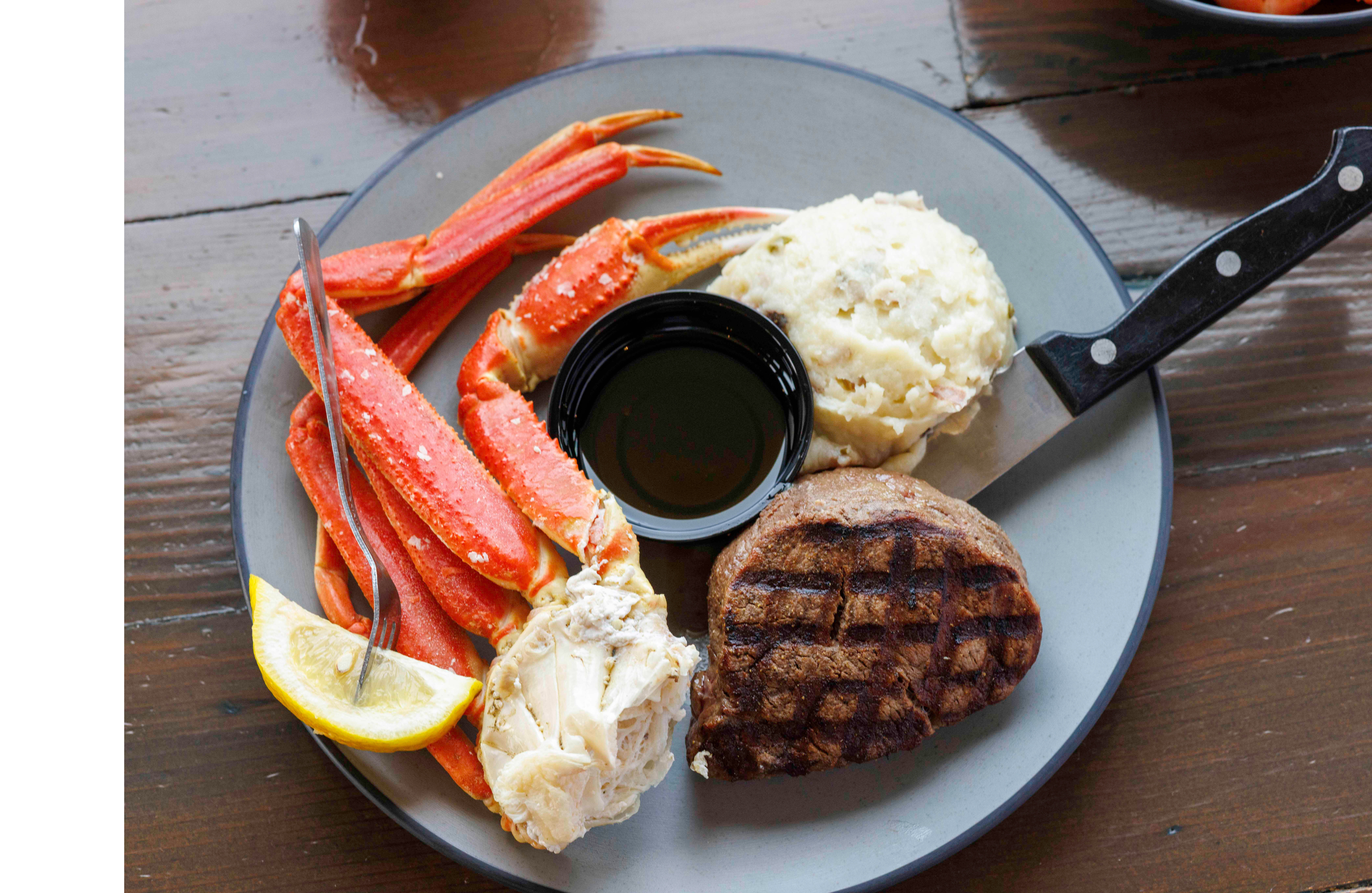 Plate with crab legs, mashed potatoes, steak, and a side of lemon on a wooden table.